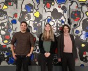 Drei Menschen stehen lächelnd vor einem großen, abstrakten Kunstwerk mit geometrischen Formen in Schwarz, Grau, Rot, Gelb und Blau. In legerer und Business-Casual-Kleidung gekleidet repräsentieren sie stolz die Hochschule Coburg mit einer Mischung aus jugendlicher Energie und Professionalität.