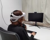 Eine Person mit langen Haaren sitzt in einem Bürostuhl und trägt ein VR-Headset. Ihr Blick geht auf einen leeren Computermonitor auf einem weißen Schreibtisch. Ihre Hand liegt auf einer Tastatur mit der Aufschrift „Hochschule Coburg“. Auf dem Monitor ist eine Webcam montiert.