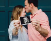 Ein Paar steht vor einer blauen Wand. Der Mann küsst die Frau auf die Stirn, während sie lächelnd ein Ultraschallbild in den Händen hält. Mit strahlender Freude feiern sie ihren bevorstehenden Nachwuchs und schwelgen in Erinnerungen an ihr erstes Treffen an der Hochschule Coburg, wo ihre gemeinsame Reise begann.