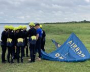 Eine Gruppe von Menschen in schwarzen Neoprenanzügen und gelben Helmen steht im Kreis neben einem Kiteboard-Segel auf einer Wiese neben einem Gewässer. Ein Mann in einem blauen Hemd, möglicherweise von der Hochschule Coburg, scheint ihnen Anweisungen zu geben. Der Himmel ist bewölkt.