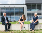 Drei Personen sitzen auf einem Betonsims vor dem modernen Gebäude der Hochschule Coburg, dessen große Fenster in der Sonne glänzen. Der Mann links trägt einen Anzug, die Frau in der Mitte ein schwarzes Oberteil und einen gestreiften Rock, und ihre Begleiterin rechts trägt ein blaues Oberteil und gestreifte Hosen. Sie lächeln und unterhalten sich.