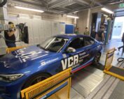 Ein blaues Auto mit den Logos „VCR“ und „Variable Compression Ratio“ steht auf einem Prüfstand in der Garage der Hochschule Coburg. Mehrere Menschen arbeiten um das Fahrzeug herum, während im Hintergrund Maschinen und Geräte geschäftiges Treiben ausmachen.