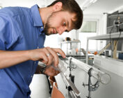 Ein Mann im blauen Hemd arbeitet fleißig in einem Labor der Hochschule Coburg und gießt Flüssigkeit in ein transparentes, auf einem Ständer montiertes Rohr. Der Raum ist mit hochmodernen wissenschaftlichen Geräten ausgestattet und hell beleuchtet.