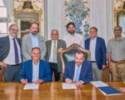 Acht Männer haben sich in einem formellen, reich verzierten Raum der Hochschule Coburg versammelt. Zwei sitzen an einem Holztisch und unterschreiben Dokumente, während sechs hinter ihnen stehen. Zur Dekoration gehören Spiegel, ein aufwendiges Wanddesign und ein großes Fenster. Alle tragen Business-Kleidung.