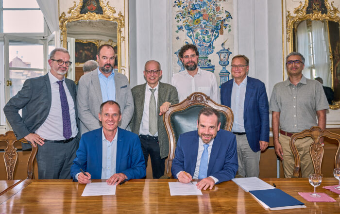 Acht Männer haben sich in einem formellen, reich verzierten Raum der Hochschule Coburg versammelt. Zwei sitzen an einem Holztisch und unterschreiben Dokumente, während sechs hinter ihnen stehen. Zur Dekoration gehören Spiegel, ein aufwendiges Wanddesign und ein großes Fenster. Alle tragen Business-Kleidung.