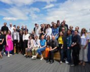 Eine Gruppe von etwa 40 Personen, die mit der Hochschule Coburg verbunden sind, posiert gemeinsam auf einem Holzdeck unter blauem Himmel. Einige sitzen auf Bänken, während andere hinter ihnen stehen. Sie tragen eine Mischung aus formeller und legerer Kleidung, rechts ist eine Backsteinmauer zu sehen.
