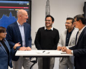 Eine Gruppe von fünf Personen in Business-Kleidung steht an einem hohen Tisch und diskutiert an der Hochschule Coburg. Im Hintergrund werden auf zwei Bildschirmen Finanzdiagramme angezeigt, was eine professionelle Atmosphäre suggeriert. Es scheint, als würden sie Ideen austauschen oder gemeinsam an einem Projekt arbeiten.