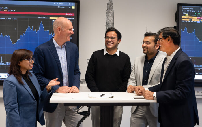 Eine Gruppe von fünf Personen in Business-Kleidung steht an einem hohen Tisch und diskutiert an der Hochschule Coburg. Im Hintergrund werden auf zwei Bildschirmen Finanzdiagramme angezeigt, was eine professionelle Atmosphäre suggeriert. Es scheint, als würden sie Ideen austauschen oder gemeinsam an einem Projekt arbeiten.
