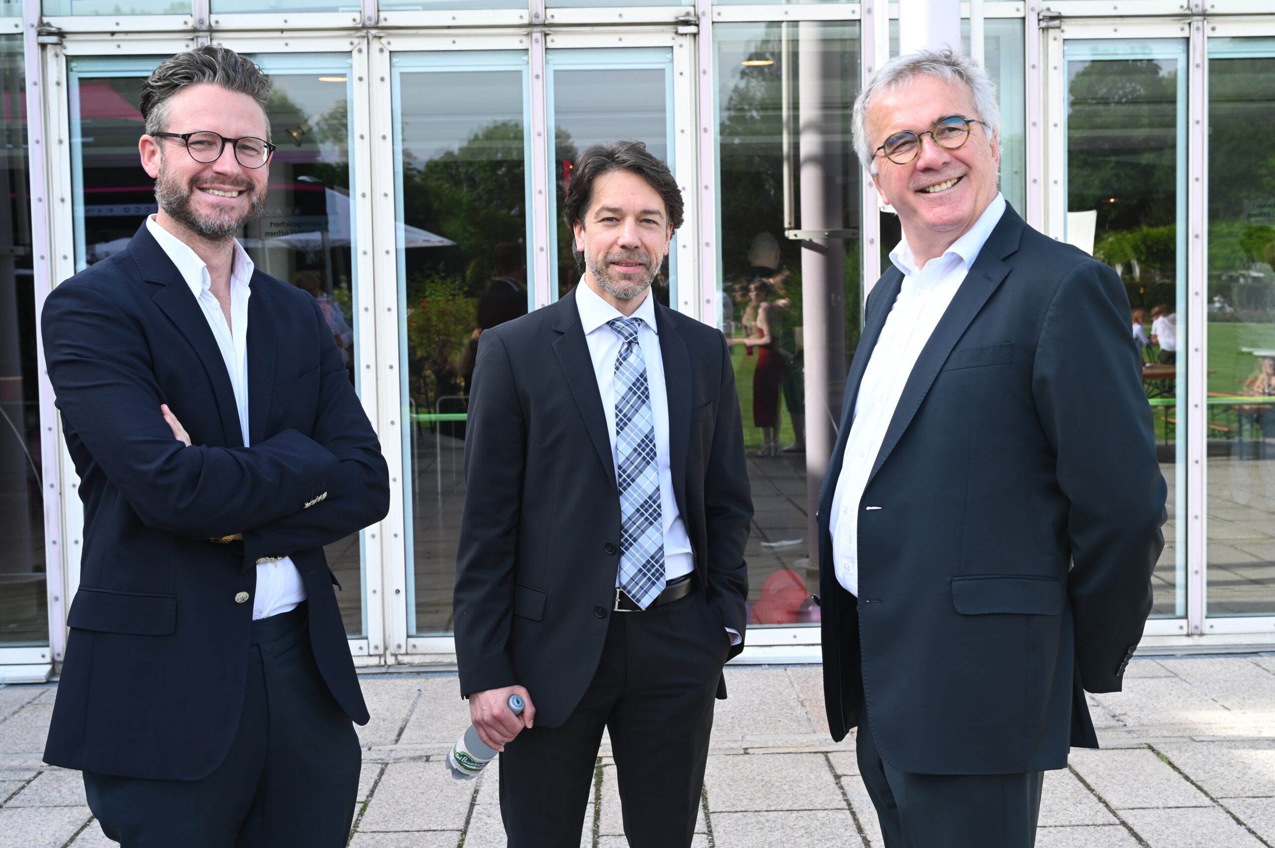 Drei Männer in Anzügen stehen vor einem modernen Gebäude mit Glasfenstern der Hochschule Coburg. Der Mann links und der Mann rechts lächeln, während der Mann in der Mitte lässig eine Wasserflasche hält. Üppige Bäume und Gras bilden eine malerische Kulisse.