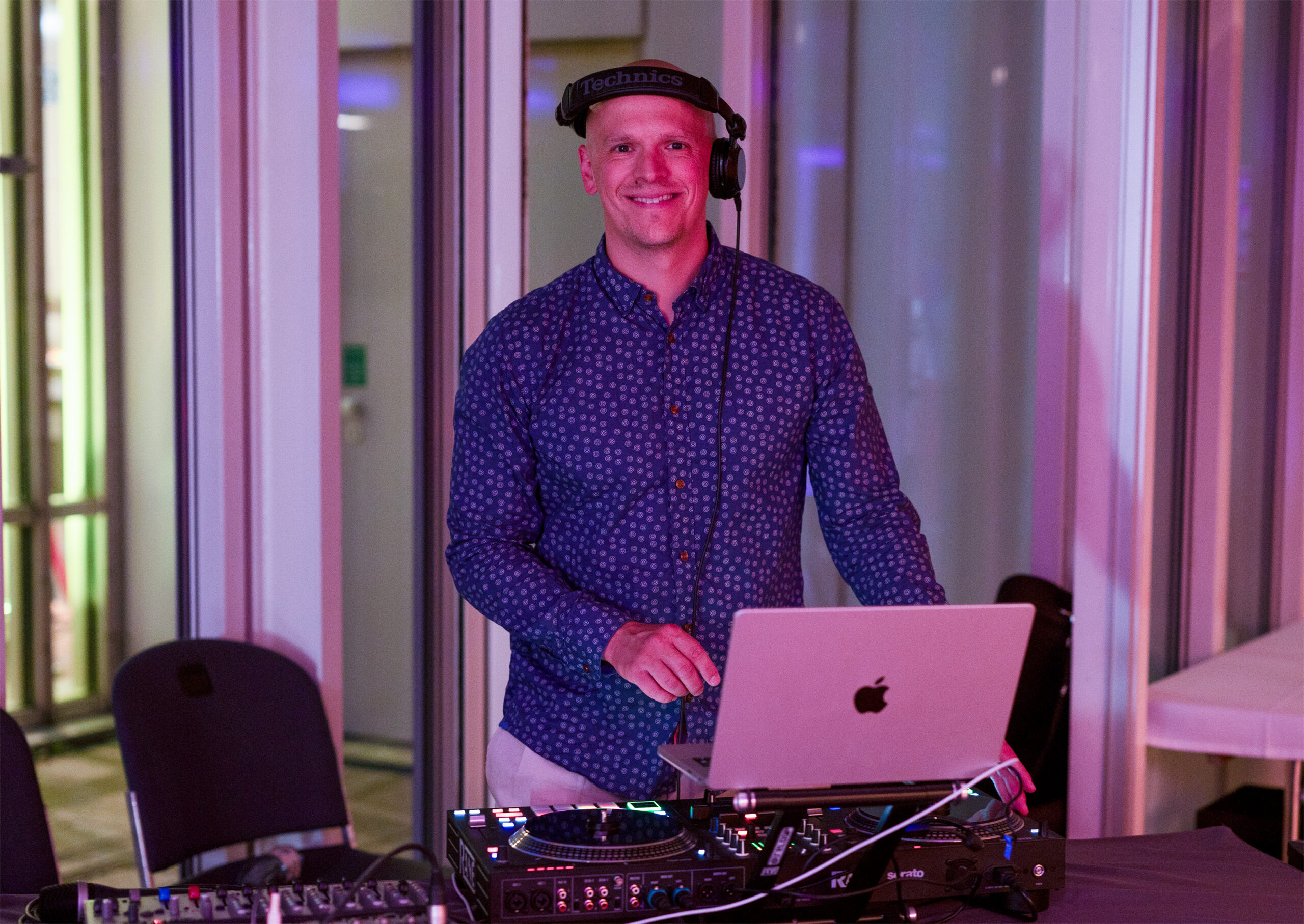 Eine Person in einem blau gemusterten Hemd und mit Kopfhörern steht bei einer Veranstaltung der Hochschule Coburg hinter einem DJ-Setup, komplett mit Laptop und Mischpult. Der Raum hat große Fenster und die Beleuchtung ist stimmungsvoll.