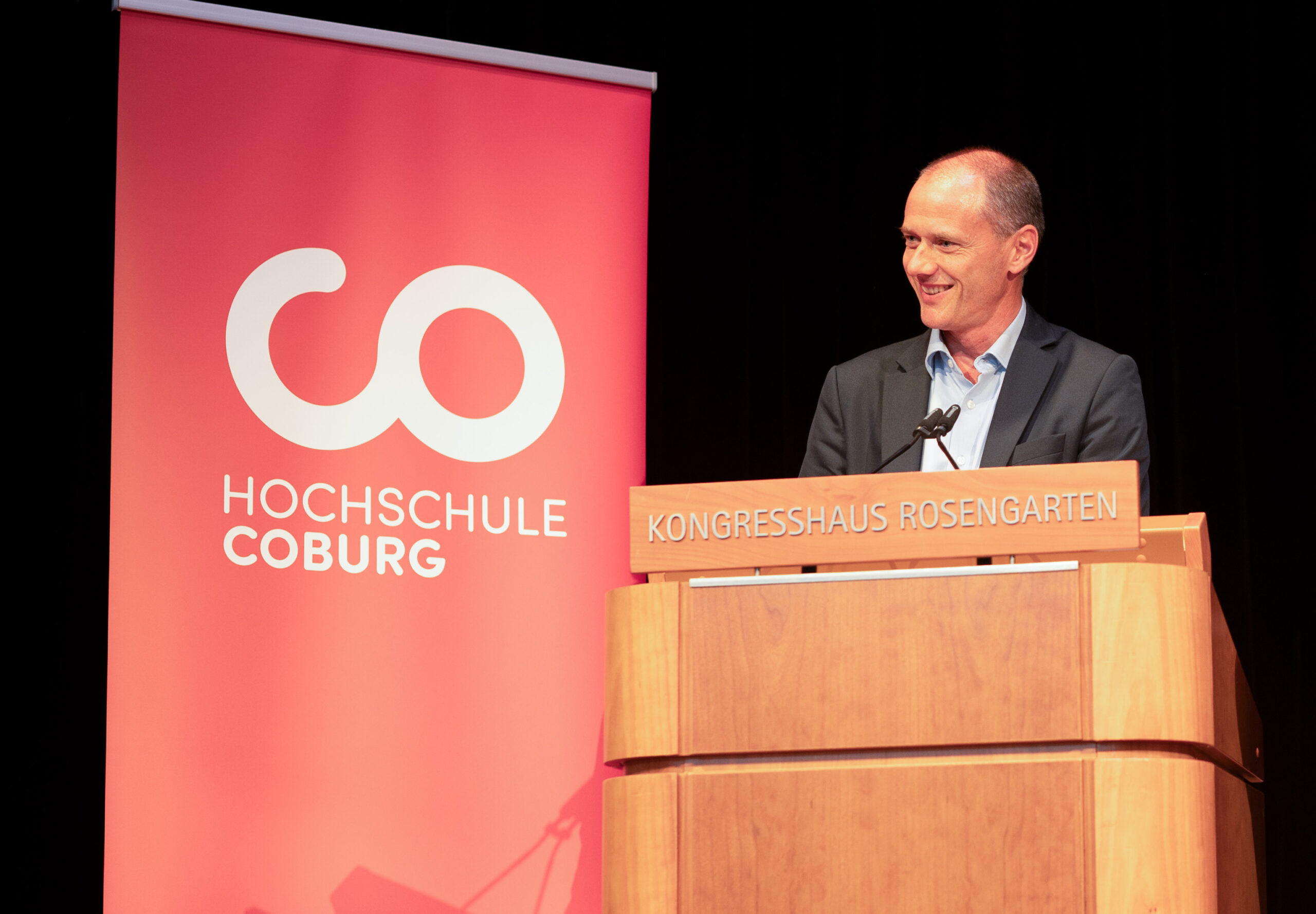 Ein Mann steht an einem Holzpodest mit der Aufschrift „Kongresshaus Rosengarten“ und spricht in ein Mikrofon. Hinter ihm prangt auf einem roten Banner stolz der Schriftzug „Hochschule Coburg“ neben einem Logo aus ineinander verschlungenen Kreisen. Er trägt einen Anzug und verkörpert damit den professionellen Geist der Hochschule Coburg.