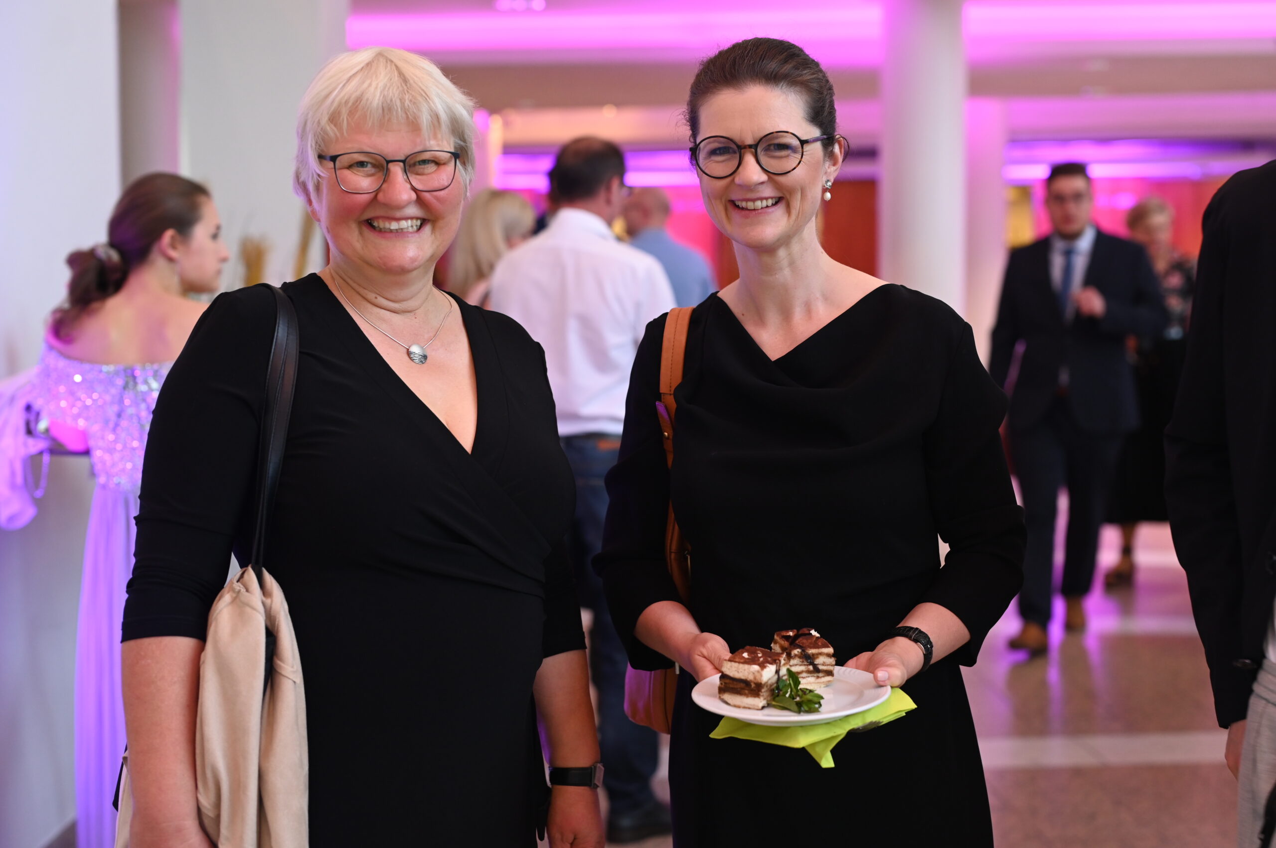 Zwei Frauen in schwarzen Kleidern lächeln bei einer Indoor-Veranstaltung an der Hochschule Coburg. Eine hält einen Teller mit Dessert. Der Hintergrund ist sanft mit rosa Licht beleuchtet und mehrere andere Personen sind anwesend und unterhalten sich.