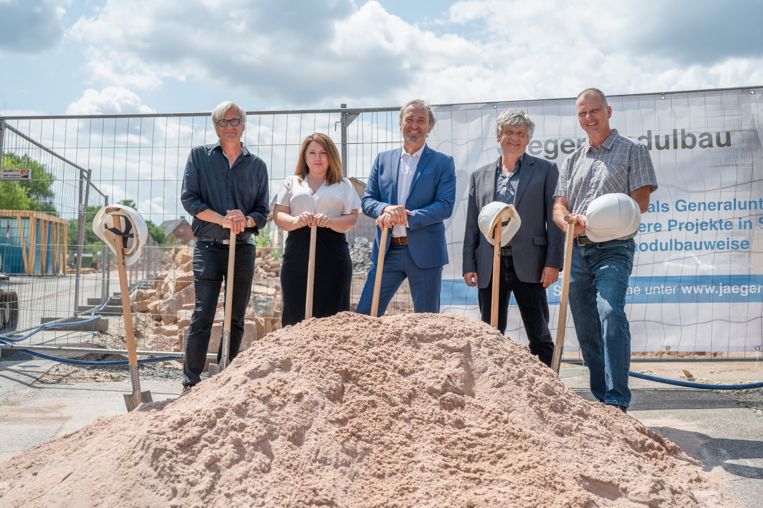 Fünf Personen in Business-Kleidung stehen mit Schaufeln in der Hand vor einer Baustelle bei der Grundsteinlegung der Hochschule Coburg. Im Hintergrund hängt stolz ein Banner mit Text, während im Vordergrund ein Sandhaufen ruht.