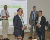 Mehrere Männer stehen in einem Konferenzraum der Hochschule Coburg. Ein Mann hält ein Geschenk in der Hand und spricht. Hinter ihnen läuft eine Präsentationsfolie. Es scheint sich um eine formelle Besprechung oder Präsentation zu handeln.