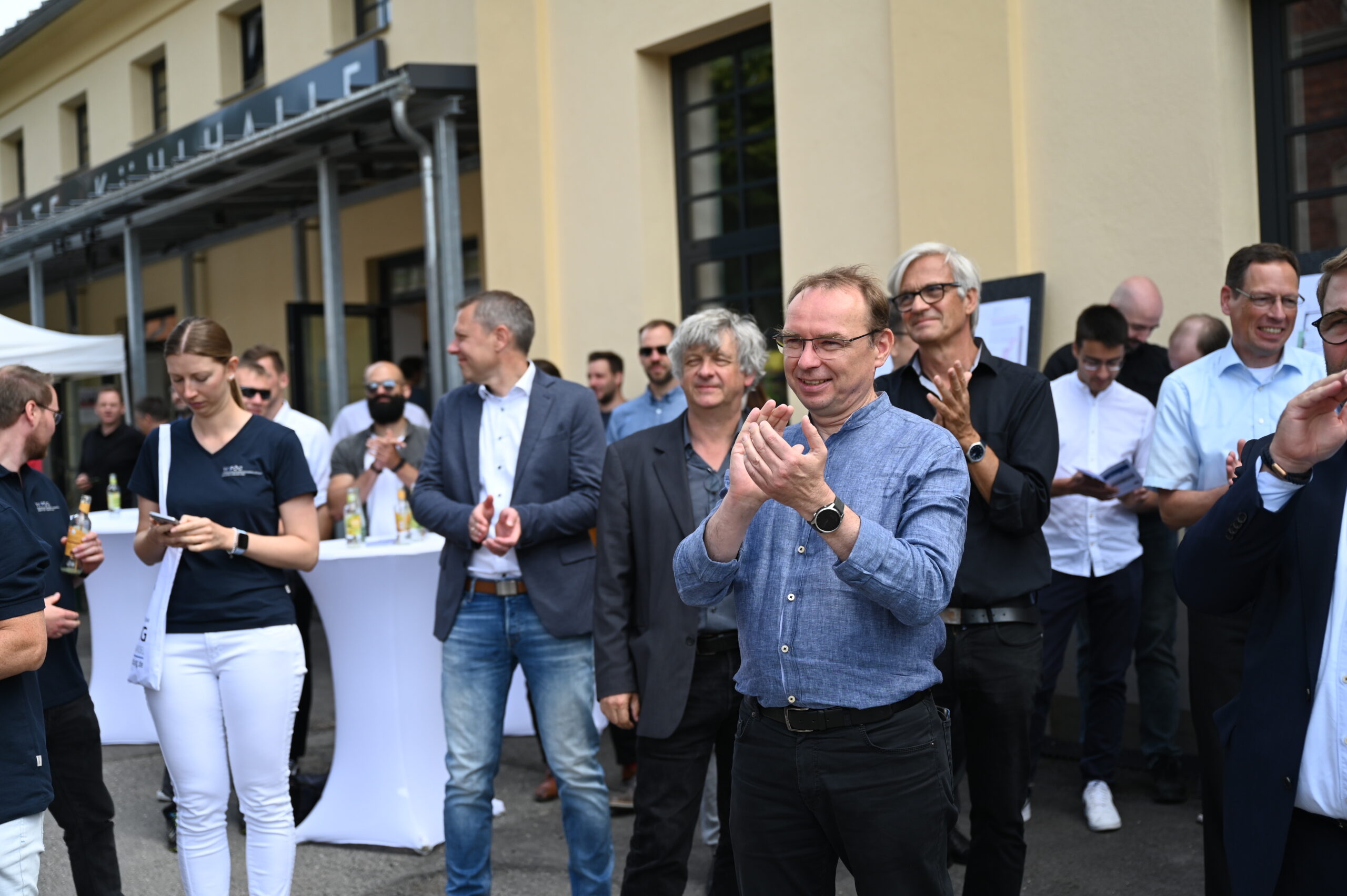 Eine Gruppe von Menschen bei einer Veranstaltung im Freien, die von der Hochschule Coburg veranstaltet wird, klatscht und lächelt. Einige sind leger gekleidet, andere haben formelle Kleidung. Im Hintergrund ist ein Buffettisch mit Getränken zu sehen und ein Gebäude mit großen Fenstern.