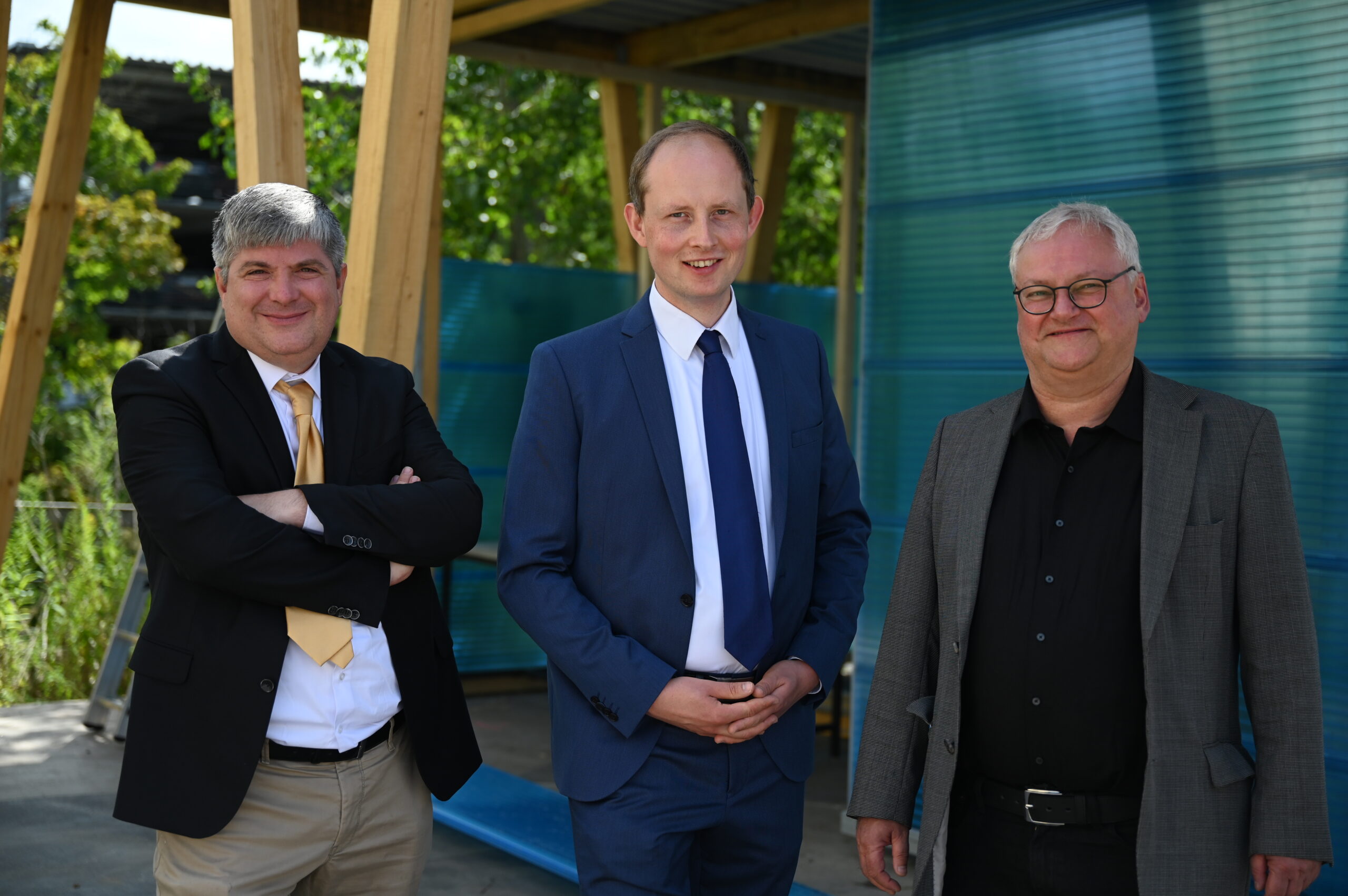 Drei Männer in Business-Kleidung stehen lächelnd im Freien. Einer trägt einen schwarzen Anzug mit gelber Krawatte, der zweite einen blauen Anzug und der dritte einen dunklen Blazer. Sie posieren selbstbewusst neben einer Holzkonstruktion der Hochschule Coburg, während üppiges Grün eine lebendige Kulisse bildet.