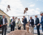 Eine Gruppe von Menschen in formeller Kleidung und mit Bauhelmen nimmt an der Grundsteinlegung für das neue Gebäude der Hochschule Coburg teil. Sie schaufeln Erde vor einer Baustelle auf, auf der vor einem festlichen Banner ein Bagger steht.