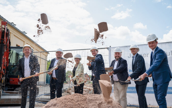 Eine Gruppe von Menschen in formeller Kleidung und mit Bauhelmen nimmt an der Grundsteinlegung für das neue Gebäude der Hochschule Coburg teil. Sie schaufeln Erde vor einer Baustelle auf, auf der vor einem festlichen Banner ein Bagger steht.