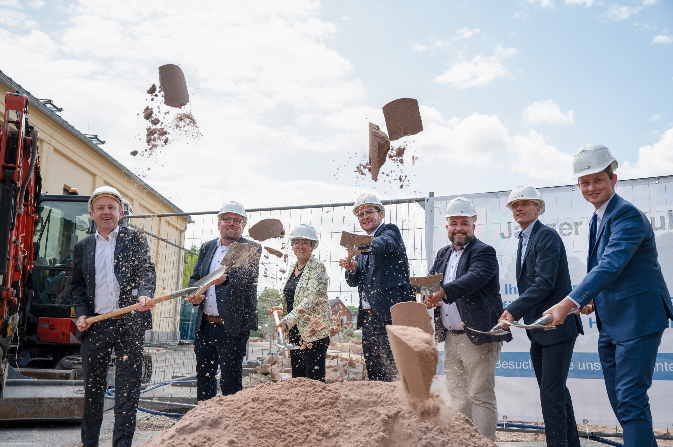 Eine Gruppe von Menschen in formeller Kleidung und mit Bauhelmen nimmt an der Grundsteinlegung für das neue Gebäude der Hochschule Coburg teil. Sie schaufeln Erde vor einer Baustelle auf, auf der vor einem festlichen Banner ein Bagger steht.