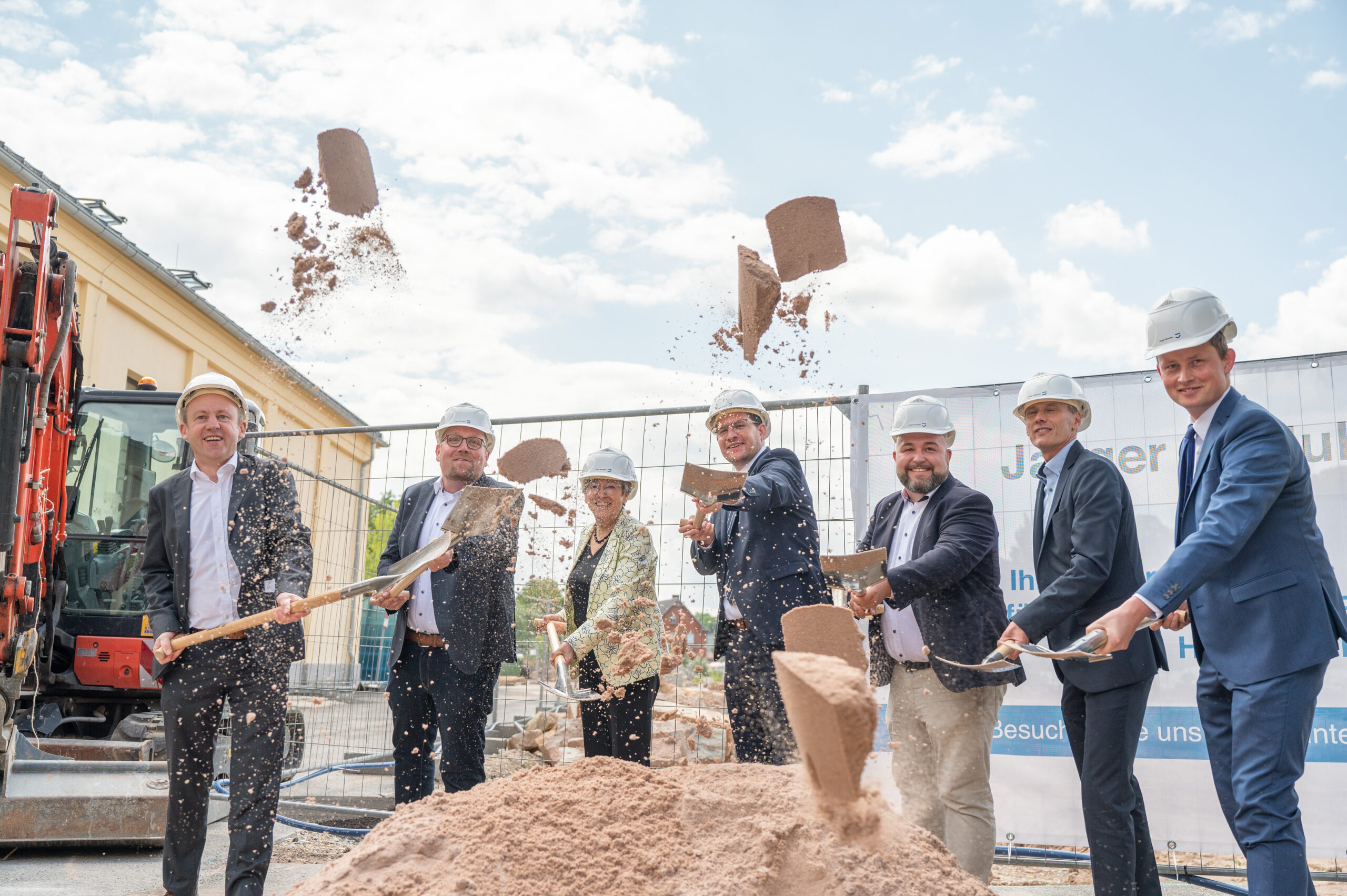 Eine Gruppe von Menschen in formeller Kleidung und mit Bauhelmen der Hochschule Coburg nimmt an einer Grundsteinlegungszeremonie teil. Jeder hält eine Schaufel in der Hand. Sie werfen Erde in die Luft. Im Hintergrund sind eine Baustelle und Maschinen zu sehen.