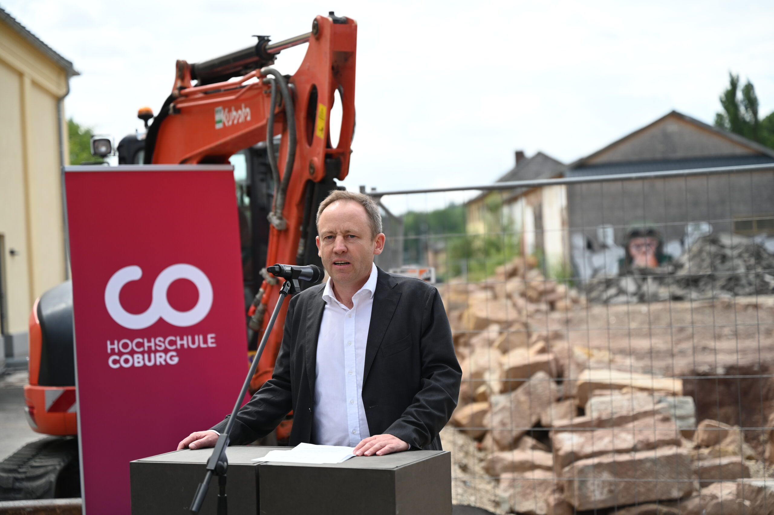 Eine Person hält eine Rede auf einem Podium, das mit einem Banner der Hochschule Coburg geschmückt ist. Dahinter arbeitet ein Bagger vor der Kulisse einer geschäftigen Baustelle, auf der Felsen und Zäune die geschäftige Landschaft prägen.