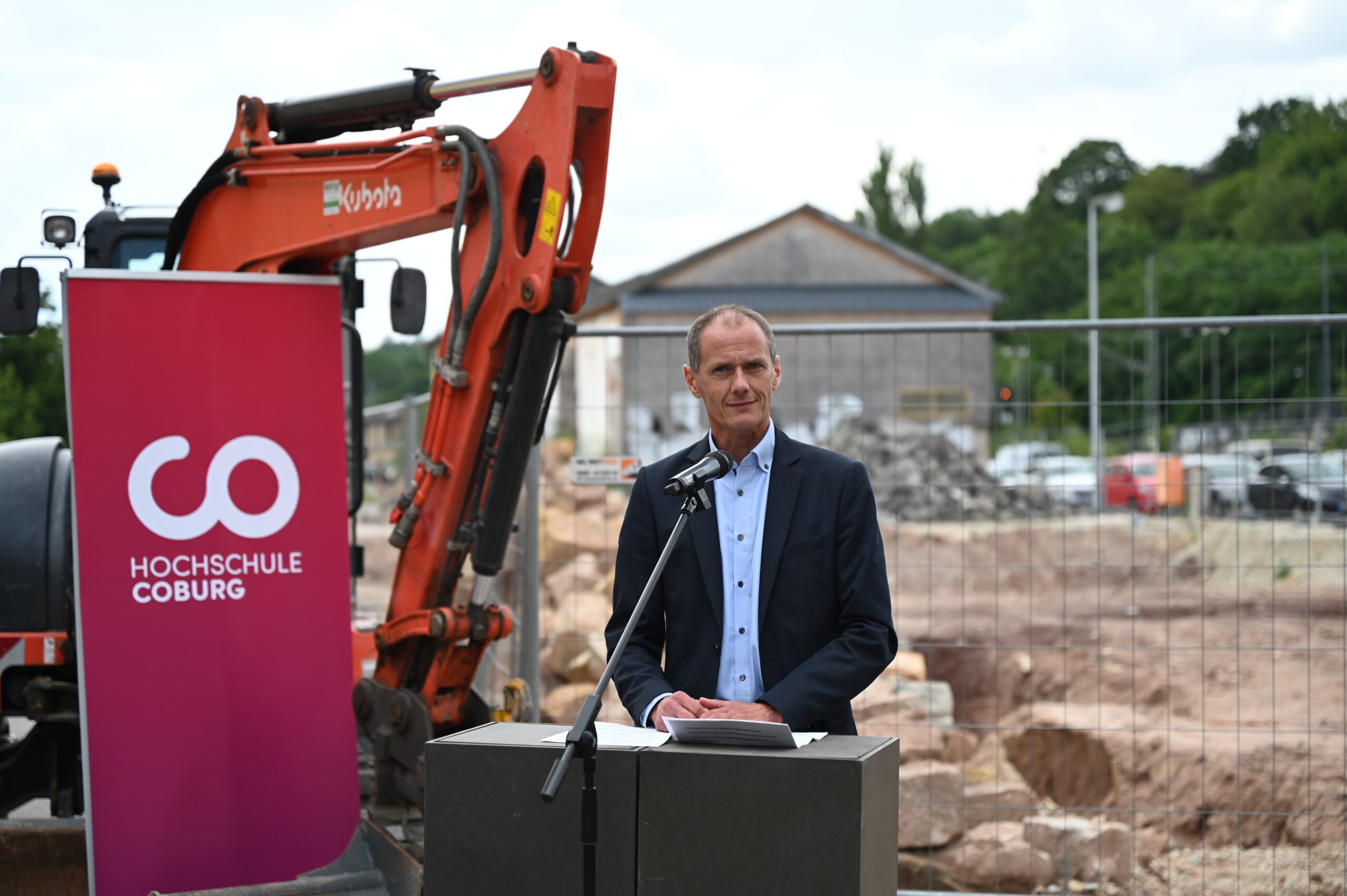 Ein Mann im Anzug spricht von einem Podium in der Nähe einer Baustelle zur Menge, auf der ein orangefarbener Bagger bereitsteht. Im Hintergrund ist das leuchtend violette Schild der Hochschule Coburg zu sehen, eingebettet zwischen Bäumen und einem nahe gelegenen Gebäude in dieser Außenkulisse.