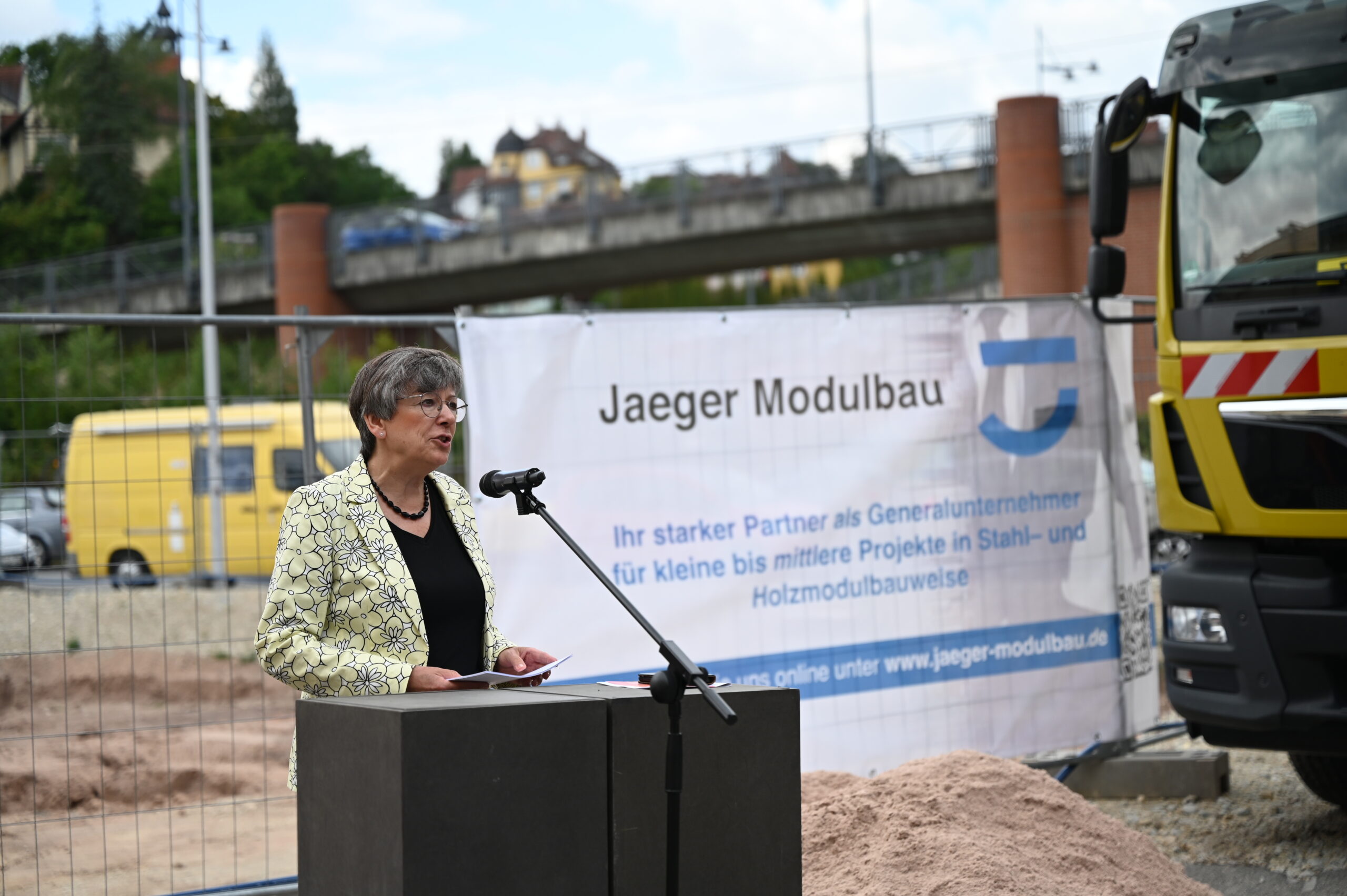Auf einer Baustelle hält eine Frau von einem Podium aus eine Ansprache an das Publikum. Hinter ihr hängt ein Banner mit der Aufschrift „Jaeger Modulbau“ und Informationen zum Modulbau auf Deutsch. In der Nähe, neben dem gelben Lieferwagen und dem Lastwagen, steht ein Hinweisschild, das vor einer Brücke und Grünflächen im Hintergrund auf die Zusammenarbeit mit der Hochschule Coburg hinweist.