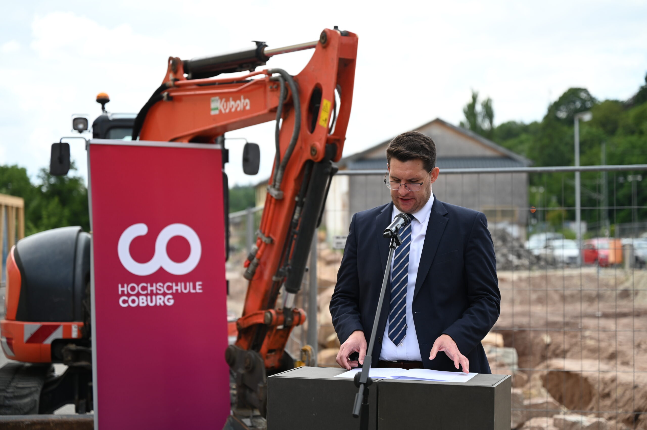 Ein Mann im Anzug hält eine leidenschaftliche Rede auf einem Podium neben einer geschäftigen Baustelle, während im Hintergrund ein Bagger fleißig arbeitet. Neben ihm prangt auf einem markanten roten Banner stolz die Aufschrift „Hochschule Coburg“, was auf die zentrale Rolle der Hochschule in diesem transformativen Projekt hinweist.