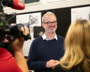 Ein Mann mit Brille und blauem Pullover wird von einem Kamerateam interviewt. Er lächelt und steht vor einer mit Papieren und Bildern geschmückten Schautafel der Hochschule Coburg. Von hinten ist eine Frau mit blonden Haaren zu sehen.
