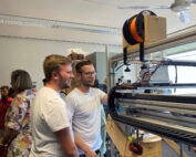 Zwei Menschen in weißen Hemden bedienen in einem Raum der Hochschule Coburg einen großen 3D-Drucker, im Hintergrund weitere. Die mit Spulen und diverser Elektronik ausgestattete Maschine schafft eine Atmosphäre, die zugleich locker und lehrreich ist.