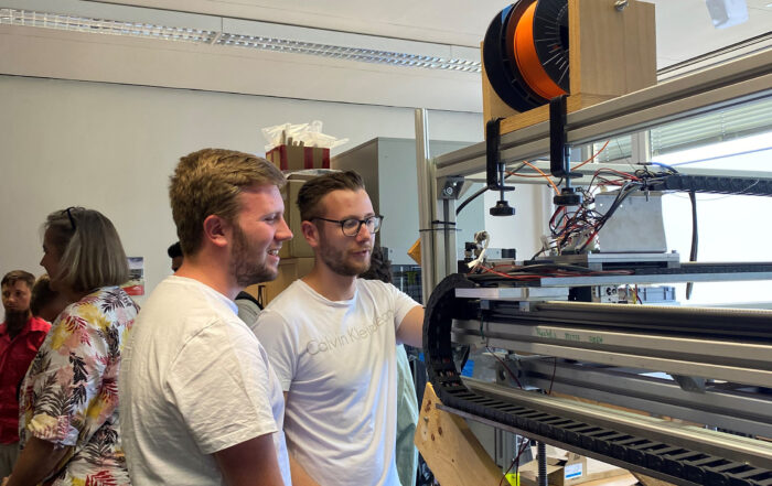 Zwei Menschen in weißen Hemden bedienen in einem Raum der Hochschule Coburg einen großen 3D-Drucker, im Hintergrund weitere. Die mit Spulen und diverser Elektronik ausgestattete Maschine schafft eine Atmosphäre, die zugleich locker und lehrreich ist.