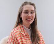 Eine Frau mit langen Haaren und Brille lächelt in die Kamera und strahlt eine Aura der Kreativität aus. Sie trägt eine farbenfrohe, gemusterte Jacke und Creolen und verkörpert damit den lebendigen Geist der Hochschule Coburg. Sie sitzt anmutig vor einem schlichten, hellen Hintergrund.