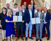 Eine Gruppe von Menschen steht auf einer mit rotem Teppich ausgelegten Treppe der Hochschule Coburg. Zwei Personen in der ersten Reihe zeigen stolz ihre Zertifikate. Sie sind alle in formeller Kleidung gekleidet und lächeln für das Foto. Im Hintergrund sind kunstvolle architektonische Details zu sehen.
