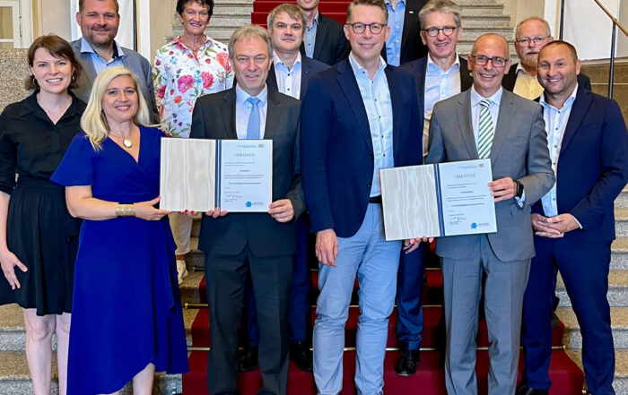 Eine Gruppe von Menschen steht auf einer mit rotem Teppich ausgelegten Treppe der Hochschule Coburg. Zwei Personen in der ersten Reihe zeigen stolz ihre Zertifikate. Sie sind alle in formeller Kleidung gekleidet und lächeln für das Foto. Im Hintergrund sind kunstvolle architektonische Details zu sehen.