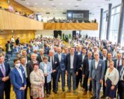 Eine große Gruppe von Menschen, darunter Männer und Frauen in formeller Kleidung, posiert für ein Gruppenfoto in einem Konferenzraum der Hochschule Coburg. Das Publikum sitzt im Hintergrund auf zwei Ebenen und die Umgebung ist hell, da natürliches Licht durch große Fenster fällt.