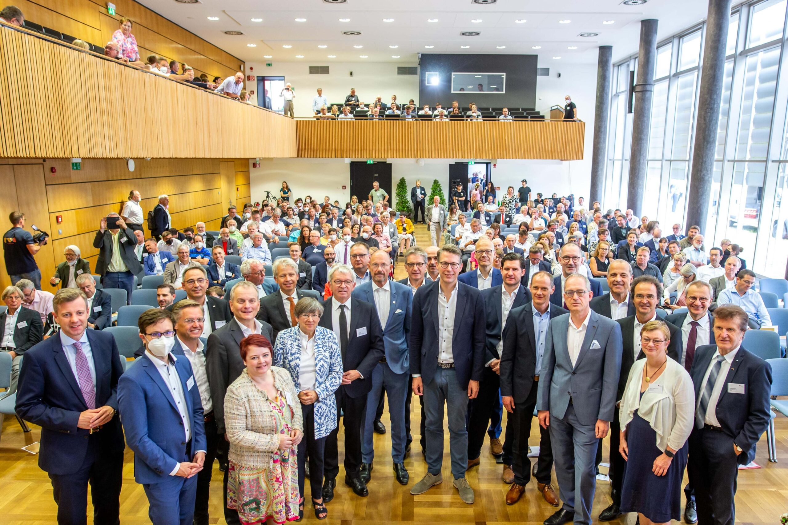 Eine große Gruppe von Menschen, darunter Männer und Frauen in formeller Kleidung, posiert für ein Gruppenfoto in einem Konferenzraum der Hochschule Coburg. Das Publikum sitzt im Hintergrund auf zwei Ebenen und die Umgebung ist hell, da natürliches Licht durch große Fenster fällt.