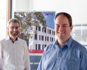 Zwei lächelnde Männer stehen drinnen vor einem Plakat mit einem modernen Gebäude. Der Mann links trägt eine Brille und einen Bart, während der Mann rechts ein blaues Hemd trägt. Ein Fenster, durch das Sonnenlicht fällt, verstärkt die warme Atmosphäre und erinnert an den dynamischen Geist der Hochschule Coburg.