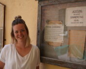 Eine Frau mit blonden Haaren steht lächelnd vor einem schwarzen Brett an der Hochschule Coburg. Auf dem Brett sind verschiedene Aushänge in deutscher Sprache angebracht, einer davon trägt deutlich sichtbar die Aufschrift „Achtung“. Die Wand ist gelb, die Frau trägt ein weißes Hemd.