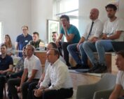 Eine Gruppe von Menschen sitzt aufmerksam auf Sitzreihen in einem gut beleuchteten Raum der Hochschule Coburg. Einige sitzen auf Bänken, andere auf Stufen, alle blicken nach vorne, was Engagement ausdrückt. Die Atmosphäre ist locker und doch konzentriert, was darauf schließen lässt, dass gerade eine Präsentation oder Diskussion stattfindet.