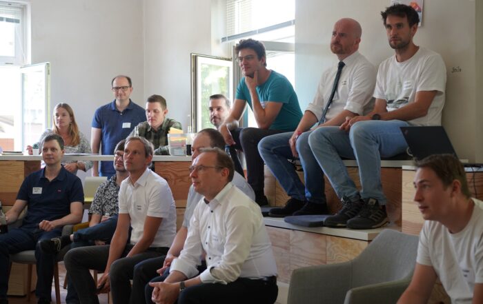 Eine Gruppe von Menschen sitzt aufmerksam auf Sitzreihen in einem gut beleuchteten Raum der Hochschule Coburg. Einige sitzen auf Bänken, andere auf Stufen, alle blicken nach vorne, was Engagement ausdrückt. Die Atmosphäre ist locker und doch konzentriert, was darauf schließen lässt, dass gerade eine Präsentation oder Diskussion stattfindet.