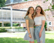 Zwei lächelnde junge Frauen stehen an einem sonnigen Tag im Freien an der Hochschule Coburg dicht beieinander. Eine trägt ein grünes Oberteil und einen geblümten Rock, während die andere ein weißes Oberteil und zerrissene Jeans trägt. Ein Backsteingebäude und ein üppiger grüner Rasen bieten die perfekte Kulisse für diesen fröhlichen Moment.