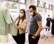 Zwei maskierte Personen untersuchen ein Kleidungsstück an einer Schaufensterpuppe in einem modernen Raum mit großen Fenstern an der Hochschule Coburg. Im Hintergrund unterhalten sich zwei weitere Personen mit Masken an einem Tisch. Tische mit Laptops und Papieren sind zu sehen und spiegeln die dynamische Bildungsumgebung wider.