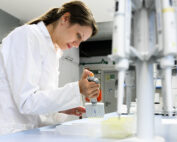 In einem Labor der Hochschule Coburg füllt ein Wissenschaftler im weißen Laborkittel mit einer Pipette Flüssigkeiten in Reagenzgläser, umgeben von hochmodernen Geräten und hellem Licht.