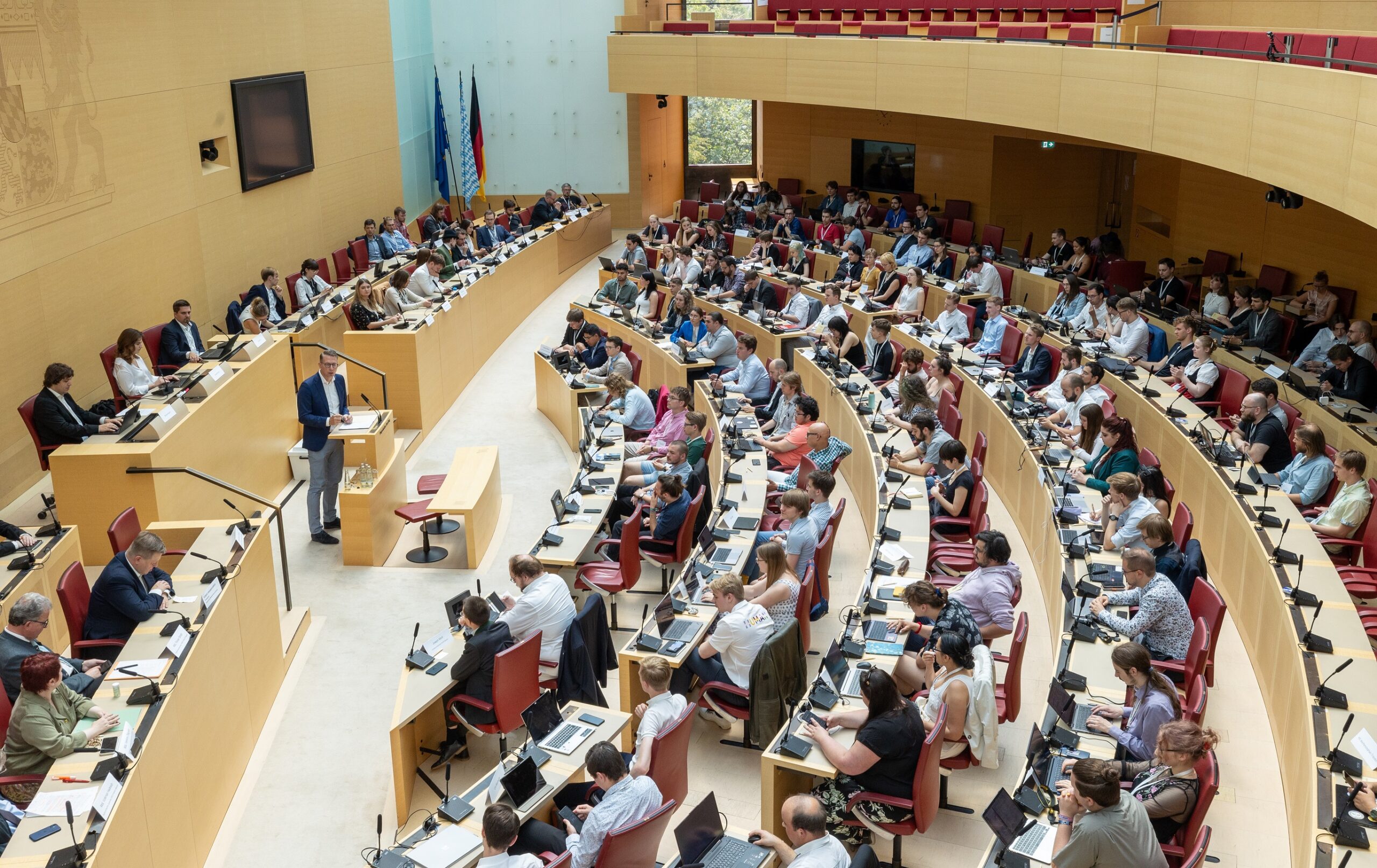 Ein großer Konferenzraum an der Hochschule Coburg ist mit Sitzreihen vollgestopft und mit Teilnehmern gefüllt. Ein Redner am Podium zieht die Aufmerksamkeit auf sich. Der gut beleuchtete Raum, ausgestattet mit einer großen Leinwand und mehreren Flaggen, erinnert an die Atmosphäre einer offiziellen Sitzung oder einer gesetzgebenden Versammlung.