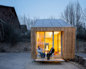 Ein kleines, modernes Holzhaus mit Solarmodulen auf dem Dach liegt in der Nähe der Hochschule Coburg. Zwei Menschen unterhalten sich auf der Veranda. Es ist Abend und das warme Licht im Inneren kontrastiert mit dem blauen Himmel. Im Hintergrund sind blattlose Bäume und ein Backsteingebäude zu sehen.