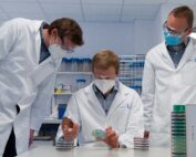 Drei Wissenschaftler in Laborkitteln und Masken untersuchen sorgfältig Petrischalen in einem Labor der Hochschule Coburg. Der Raum ist voller Regale und hochmoderner Geräte, auf dem Tisch liegen Stapel von Petrischalen.