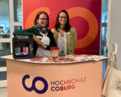 Zwei Frauen stehen hinter einem Stand der Hochschule Coburg, an dem Broschüren, rote Stifte und andere Materialien ordentlich angeordnet sind. Ein 3D-Drucker auf der linken Seite des Tisches sorgt für einen modernen Touch. Im Hintergrund ist auf einem Banner das Logo der Hochschule Coburg prominent zu sehen.