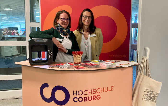 Zwei Frauen stehen hinter einem Stand der Hochschule Coburg, an dem Broschüren, rote Stifte und andere Materialien ordentlich angeordnet sind. Ein 3D-Drucker auf der linken Seite des Tisches sorgt für einen modernen Touch. Im Hintergrund ist auf einem Banner das Logo der Hochschule Coburg prominent zu sehen.
