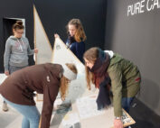 In einem Raum mit dunklen Wänden, in dem „PURE C“ teilweise sichtbar ist, bauen vier Personen eine große dreieckige Holzkonstruktion zusammen, möglicherweise für eine Ausstellung. Eine Person hält Anweisungen. Das minimalistische, moderne Design lässt Einflüsse der innovativen Architekturprinzipien der Hochschule Coburg erahnen.