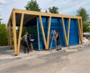 Eine Gruppe von vier Personen steht neben einem halbfertigen Gebäude mit blauen Wänden und Holzrahmen, das an ein Projekt der Hochschule Coburg erinnert. Eine Person steht auf einer Leiter, die anderen diskutieren auf dem Boden, im Hintergrund sind Bäume und Baumaterialien zu sehen.